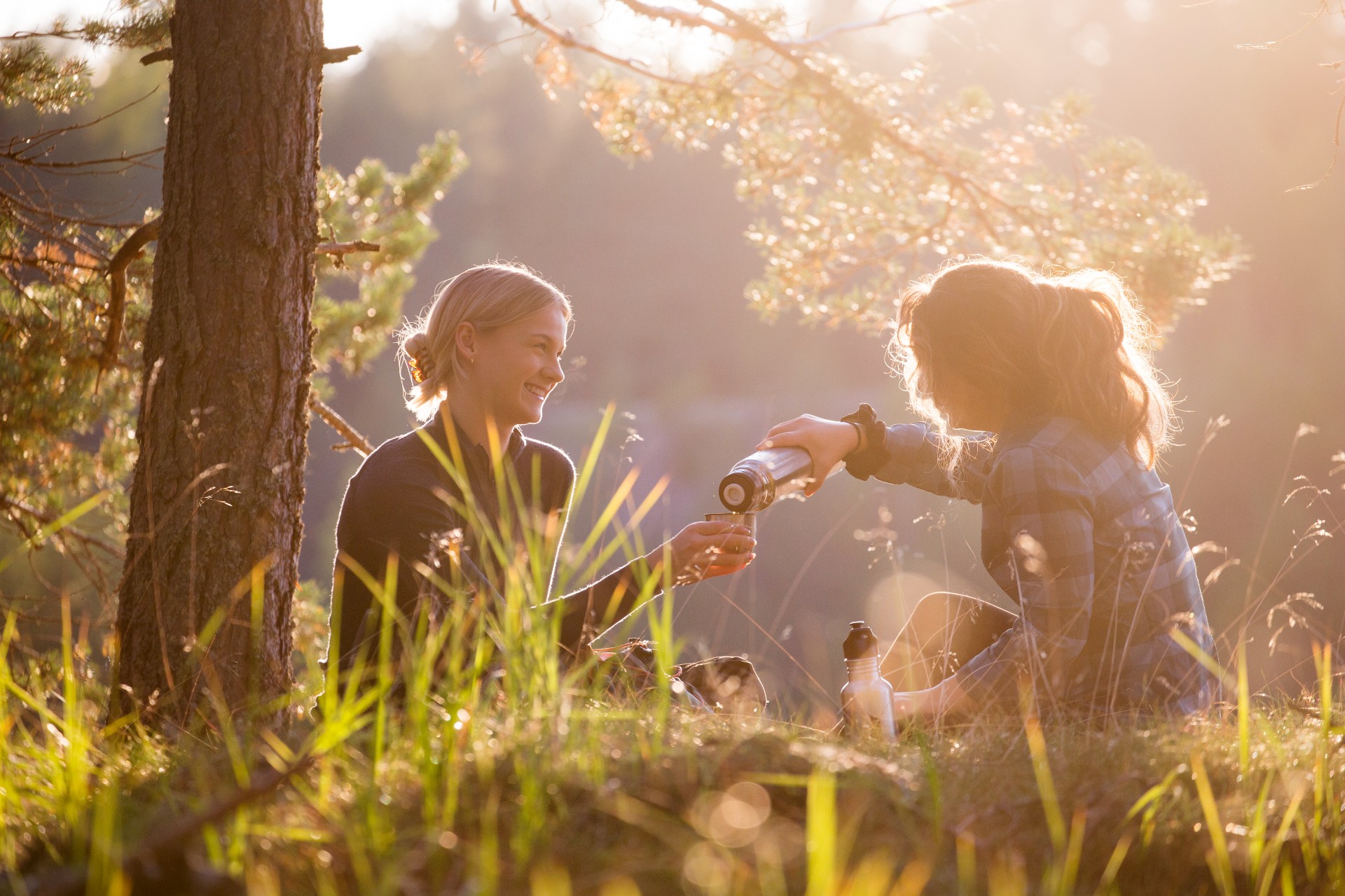 Two women sitting in a sunny forest, drinking coffee.